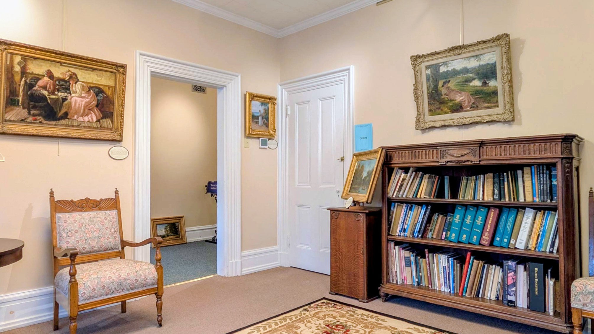 Gallery corner hung with gilt-framed 19th-century oil paintings above a bookcase of art volumes and a Victorian side chair.