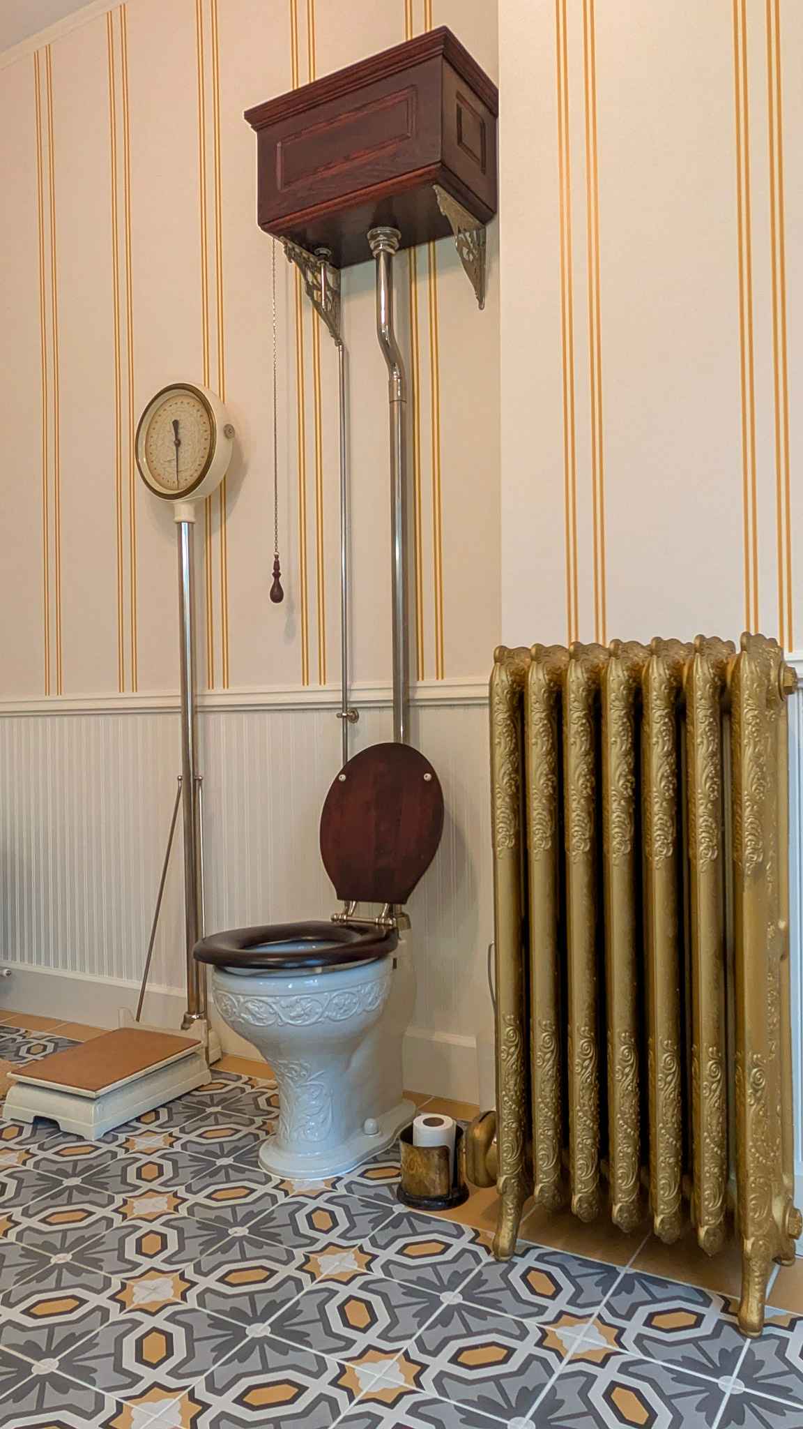 Restored Victorian water closet with a high wooden pull-chain tank, gilded cast-iron radiator, and encaustic tile floor.