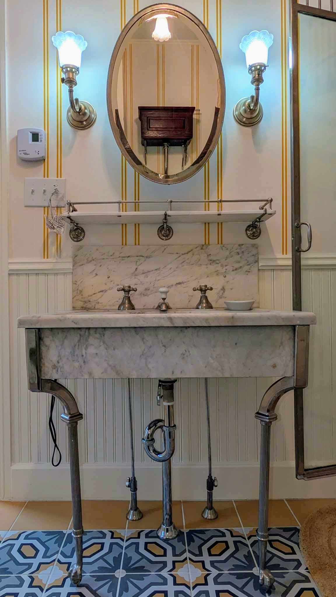 Marble console sink on nickel legs below an oval mirror and blue glass sconces in a tiled Victorian bath.