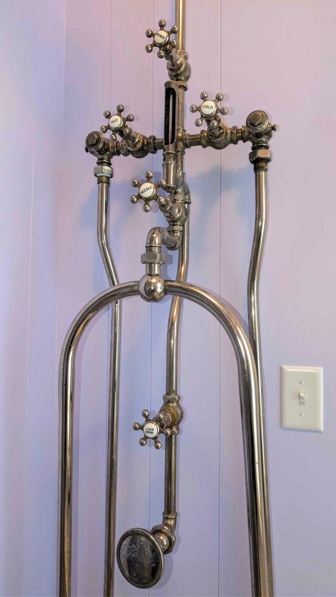 Exposed nickel shower and tub plumbing with cross-handle valves against a lavender wall.