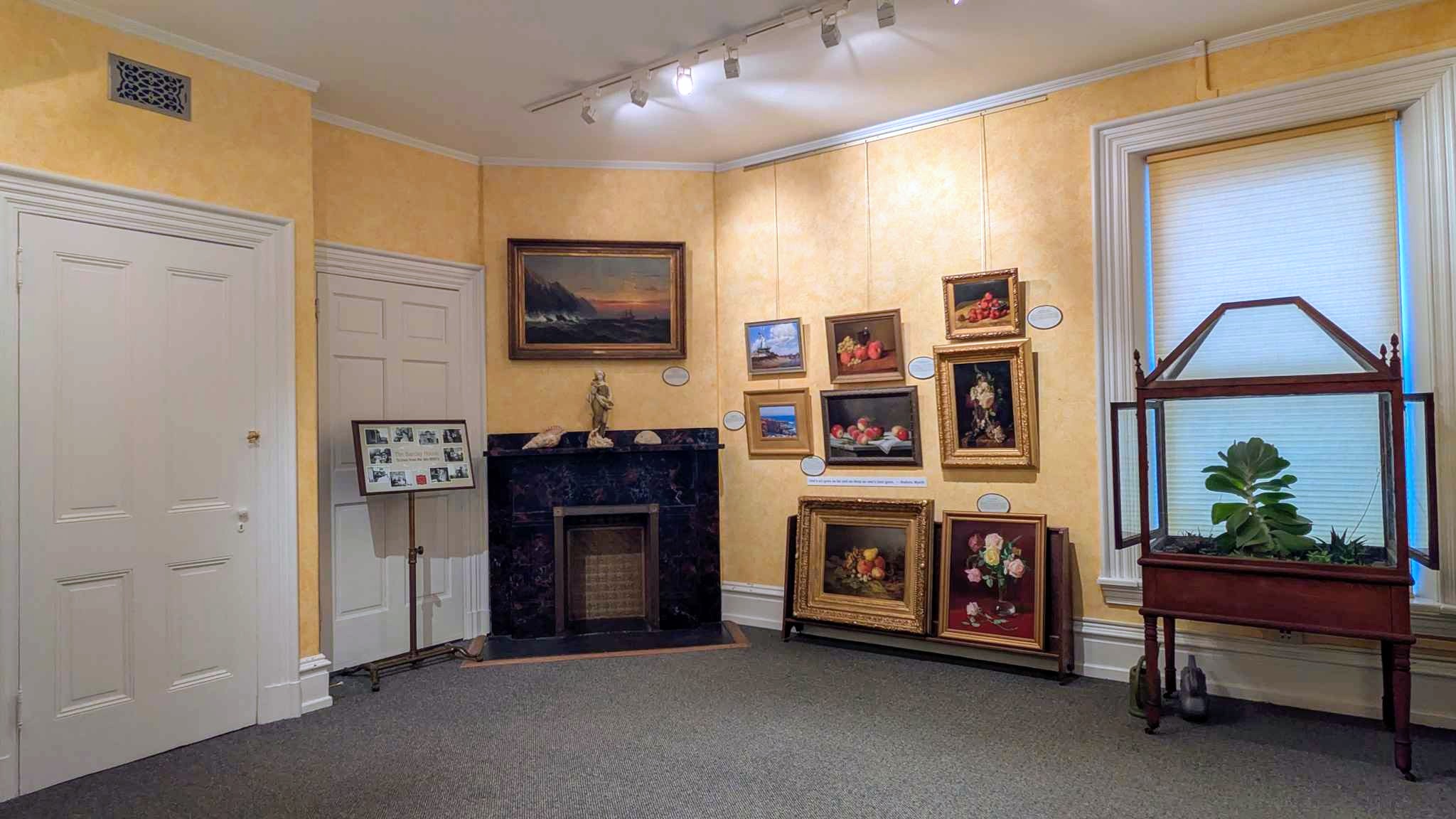 Yellow gallery room with a black marble mantel and salon-hung 19th-century still lifes and landscapes.