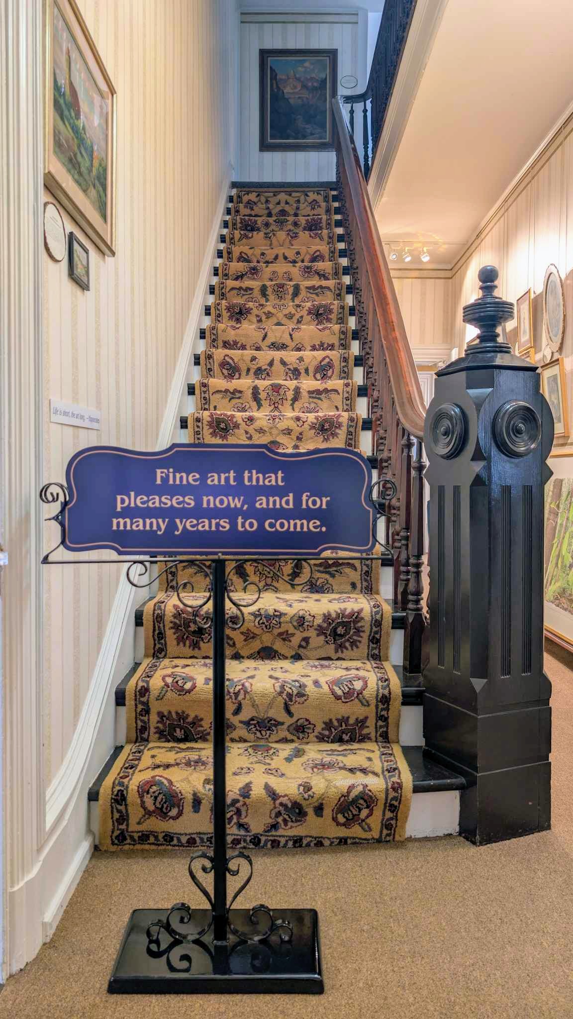 Staircase with a floral runner beside a gallery sign reading "Fine art that pleases now, and for many years to come."