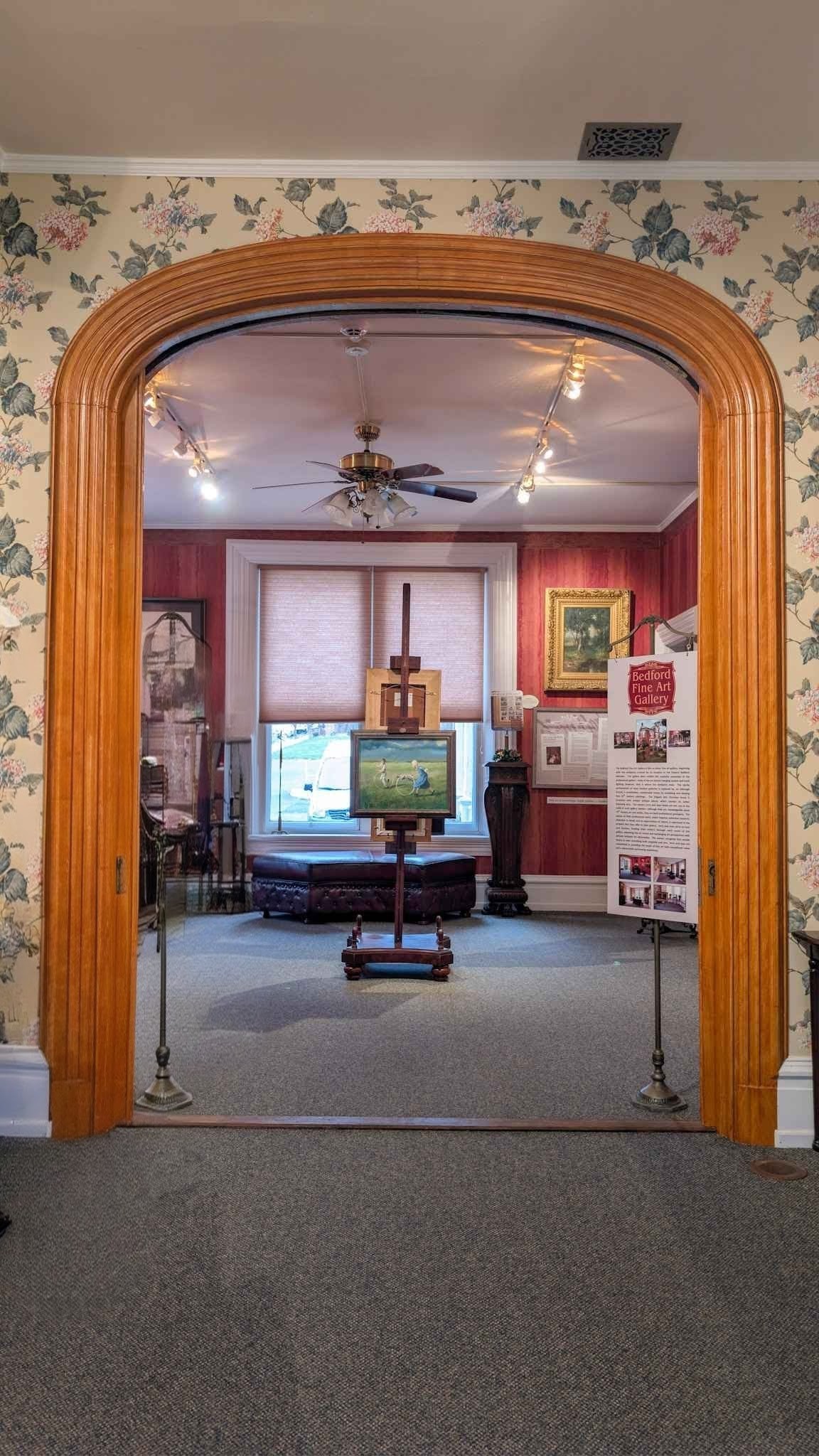 View through a carved oak archway into a red gallery room with a painting displayed on an easel.