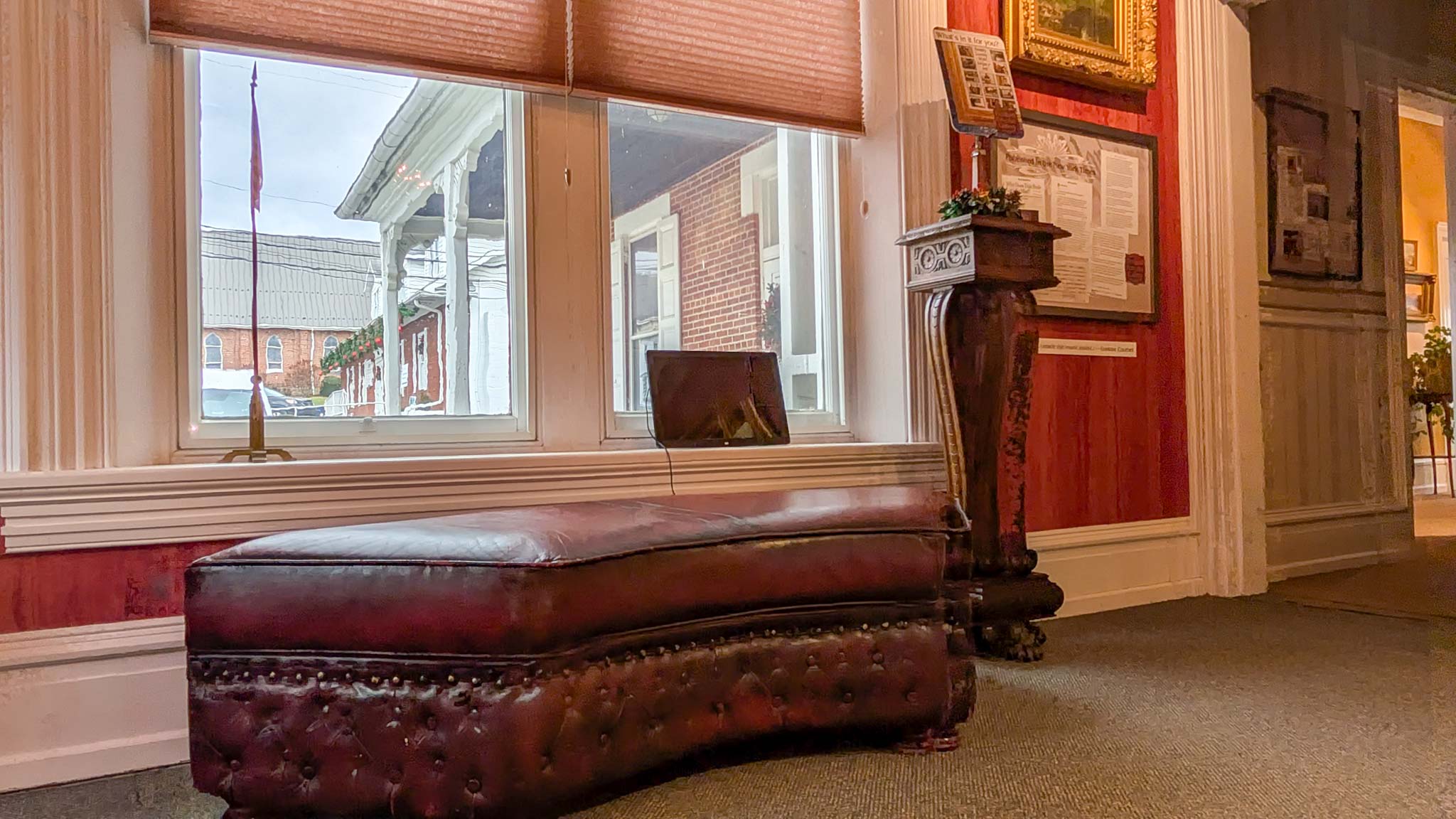 Tufted burgundy leather chaise beneath a window overlooking Bedford's streetscape in a red gallery room.