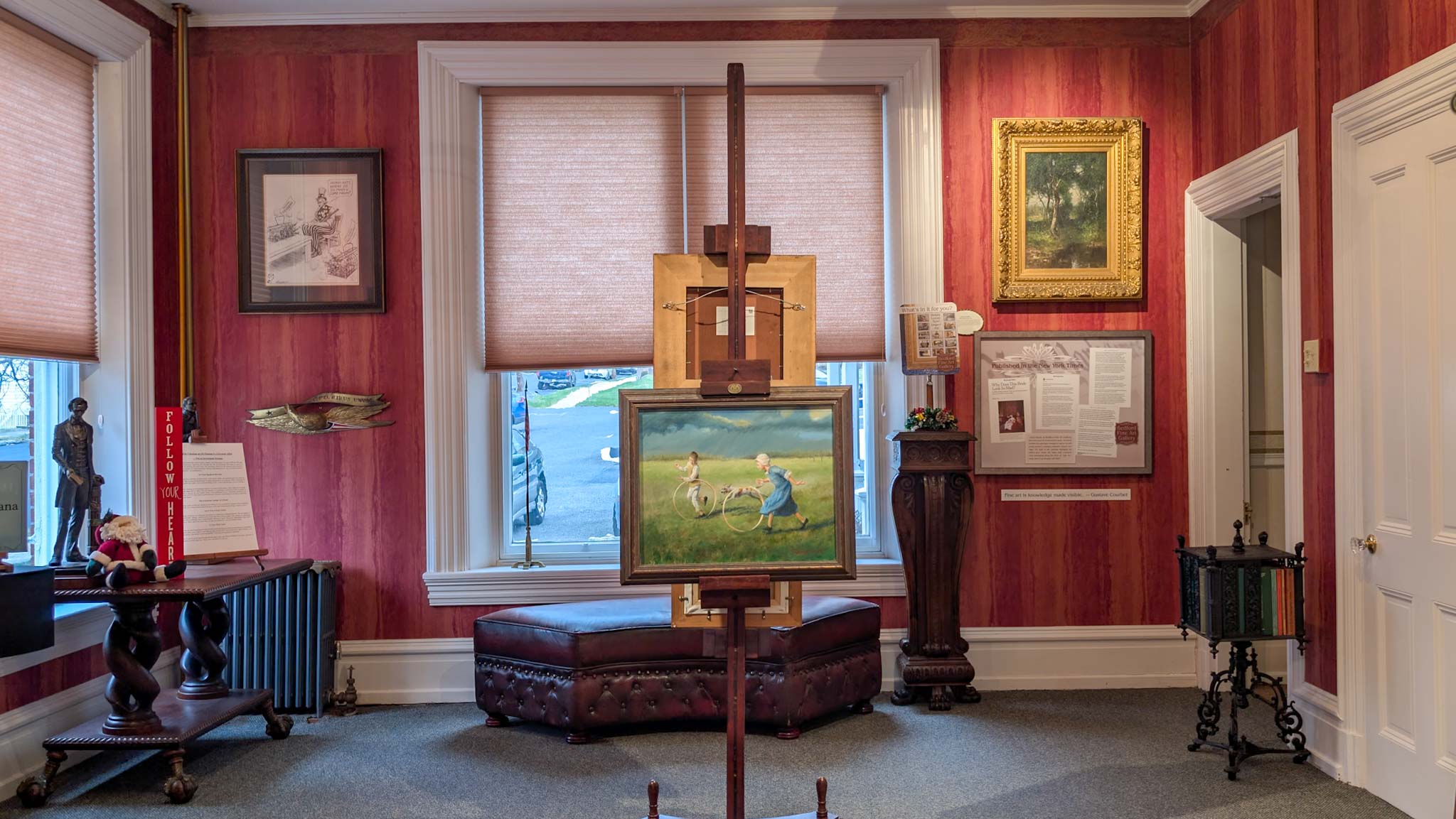 Red gallery room with a landscape painting on an easel between framed artworks and a cast-iron radiator.