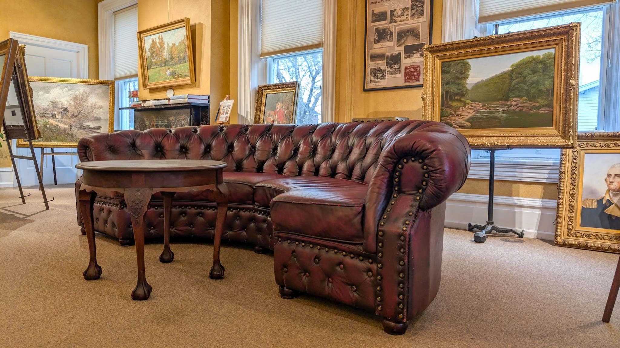 Curved tufted oxblood leather sofa amid easels of framed paintings in a sunlit gallery room.