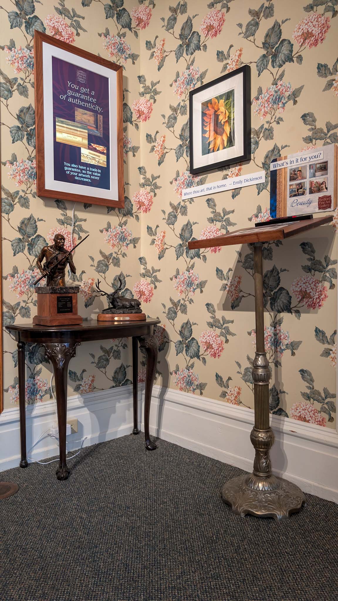 Floral-papered corner with a bronze sculpture on a console table and framed gallery signage.