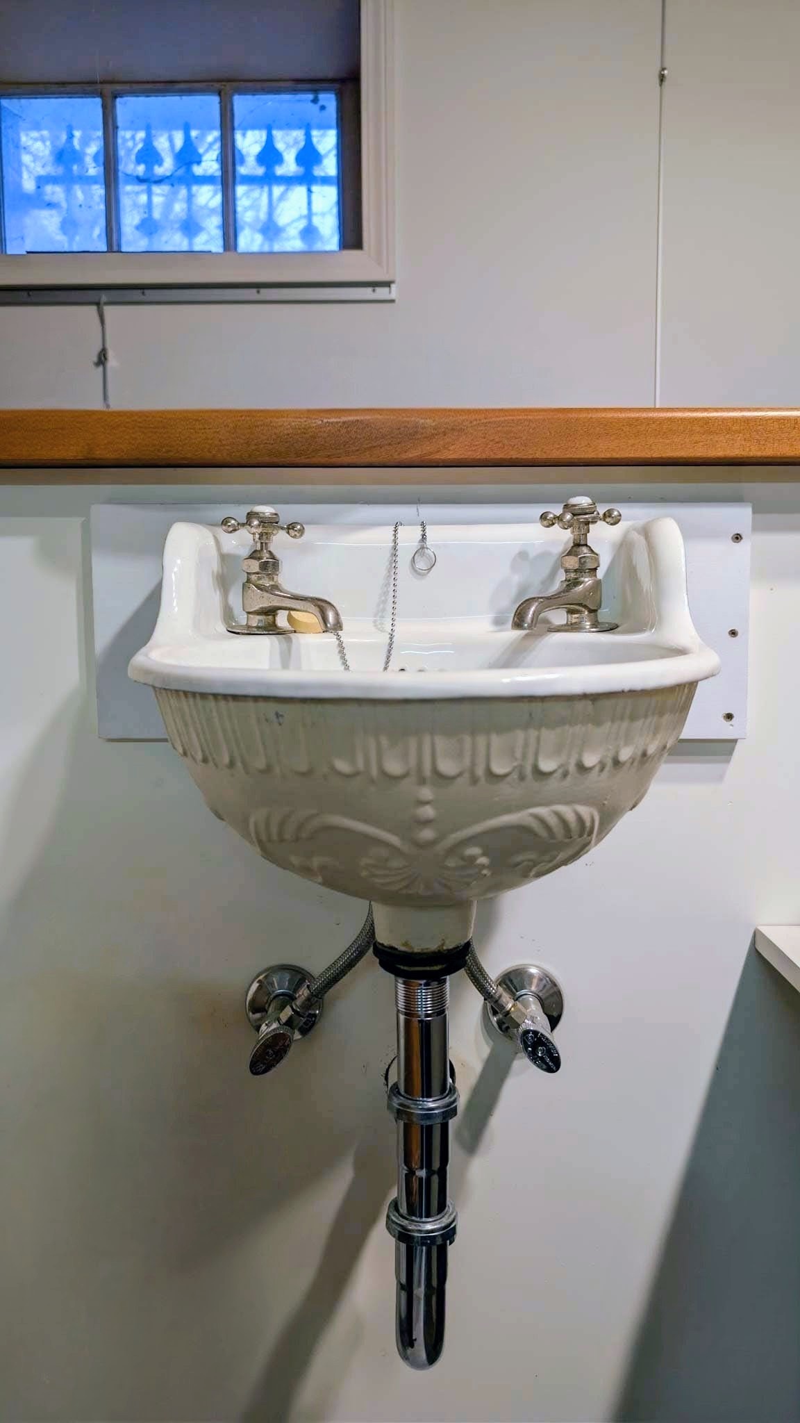 Wall-mounted embossed ironstone sink with cross-handle taps below a stained-glass window.