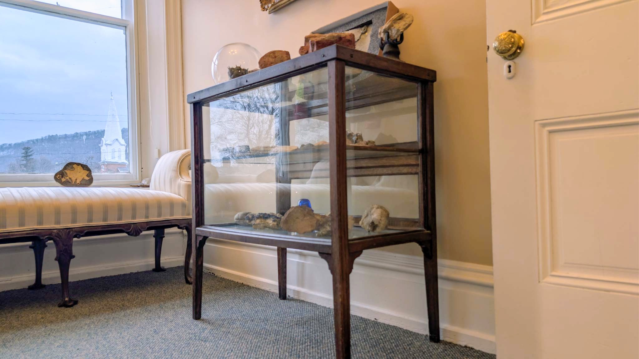 Glass specimen cabinet of minerals beside a window bench, a church steeple visible outside.