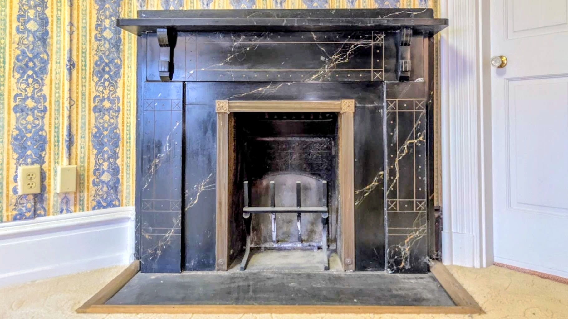Black marbleized fireplace with gilt detailing against blue floral Victorian wallpaper.